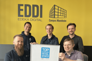 Vor der gelben Hintergrundwand mit dem Eddi Logo von Edeka sitzen im Kreis Dominik, Laszlo, Patrick, Thomas und Jens und halten die "Top Supplier" Urkunde.